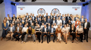 Das Bild zeigt eine große Gruppe von Menschen in festlicher Kleidung, die in mehreren Reihen vor einer Bühne posieren. Im Hintergrund ist eine große Leinwand mit Logos des Deutschen Instituts für Service-Qualität und von n-tv zu sehen sowie dem Schriftzug „Deutschlands Beste Online-Portale 2025“. Die Anwesenden halten Trophäen in der Hand – es handelt sich um eine Preisverleihung.